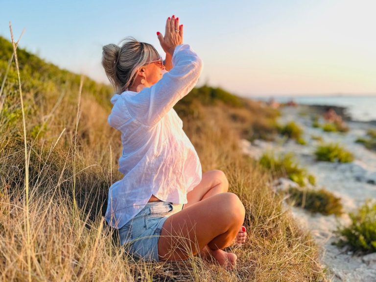 Frau sitzt meditierend am Strand, mit erhobenen Händen und Sonnenuntergang im Hintergrund.