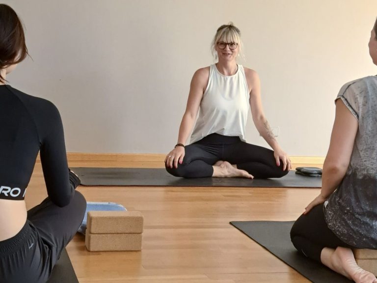 Drei Personen sitzen in einer Yoga-Position, während eine Lehrerin Anleitung gibt.