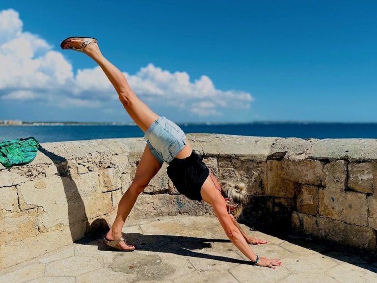 Frau in Sportkleidung macht eine Yoga-Übung am Meer unter blauem Himmel.